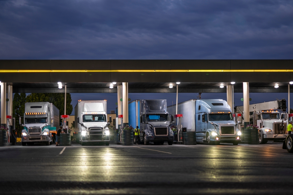 Industrial carriers big rig semi trucks with semi trailers fill the tanks with diesel fuel standing on the truck stop gas station with lights at night to be ready to continue the route by morning