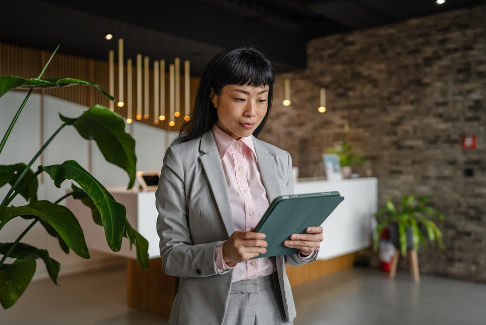 Asian businesswoman in grey suit and pink shirt holds green tablet in a modern lobby by reception and plants, conveying professional tech-savvy leadership and corporate connectivity
